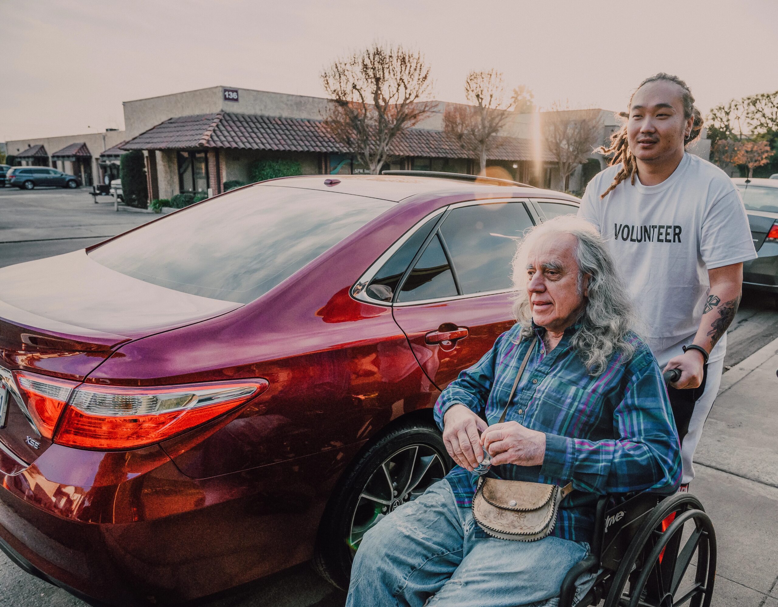 fun activities for dogs while owners are at work - A volunteer helps an elderly man in a wheelchair near parked cars on a sunny day.
