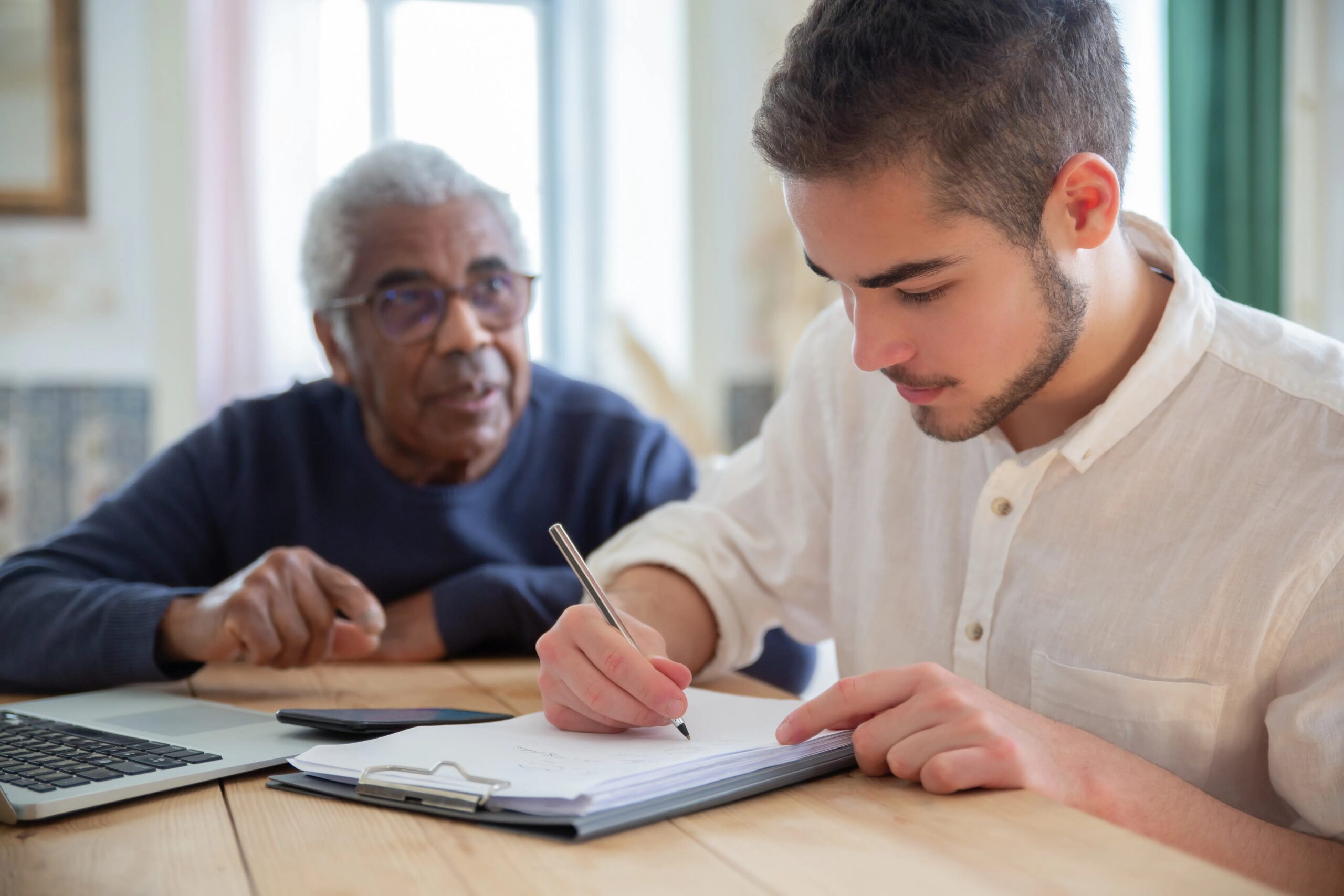 benefits of professional dog training classes - A young man aids a senior citizen with writing at a wooden table indoors, fostering social assistance and connection.